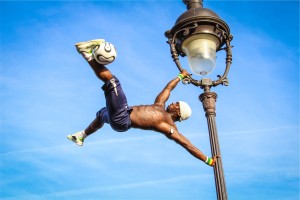 man holding onto light pole horizontally kicking soccer ball